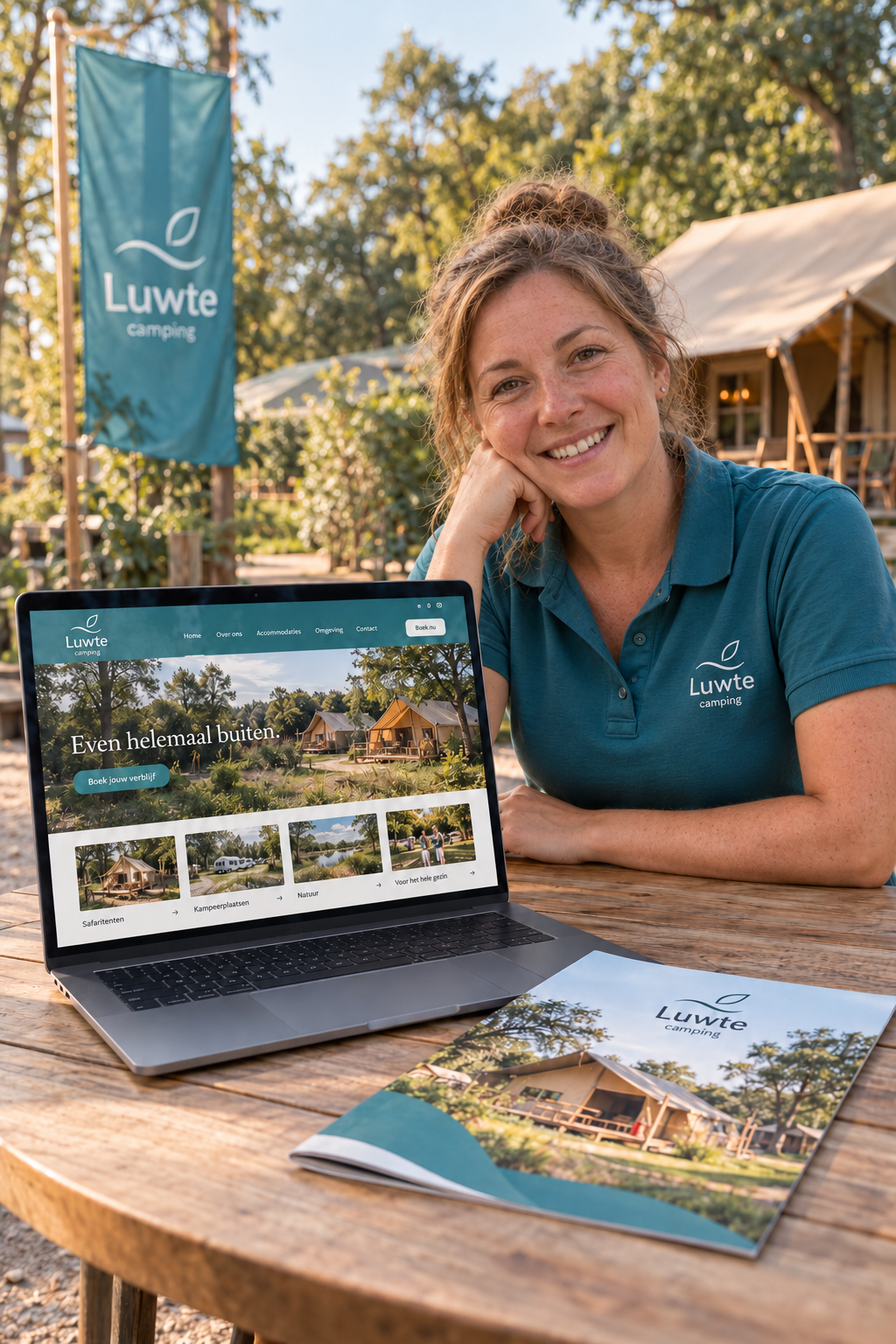 A smiling campsite host in a teal polo shirt sits at an outdoor table with a laptop showing her campsite's website, a brochure beside it and safari tents among the trees behind her